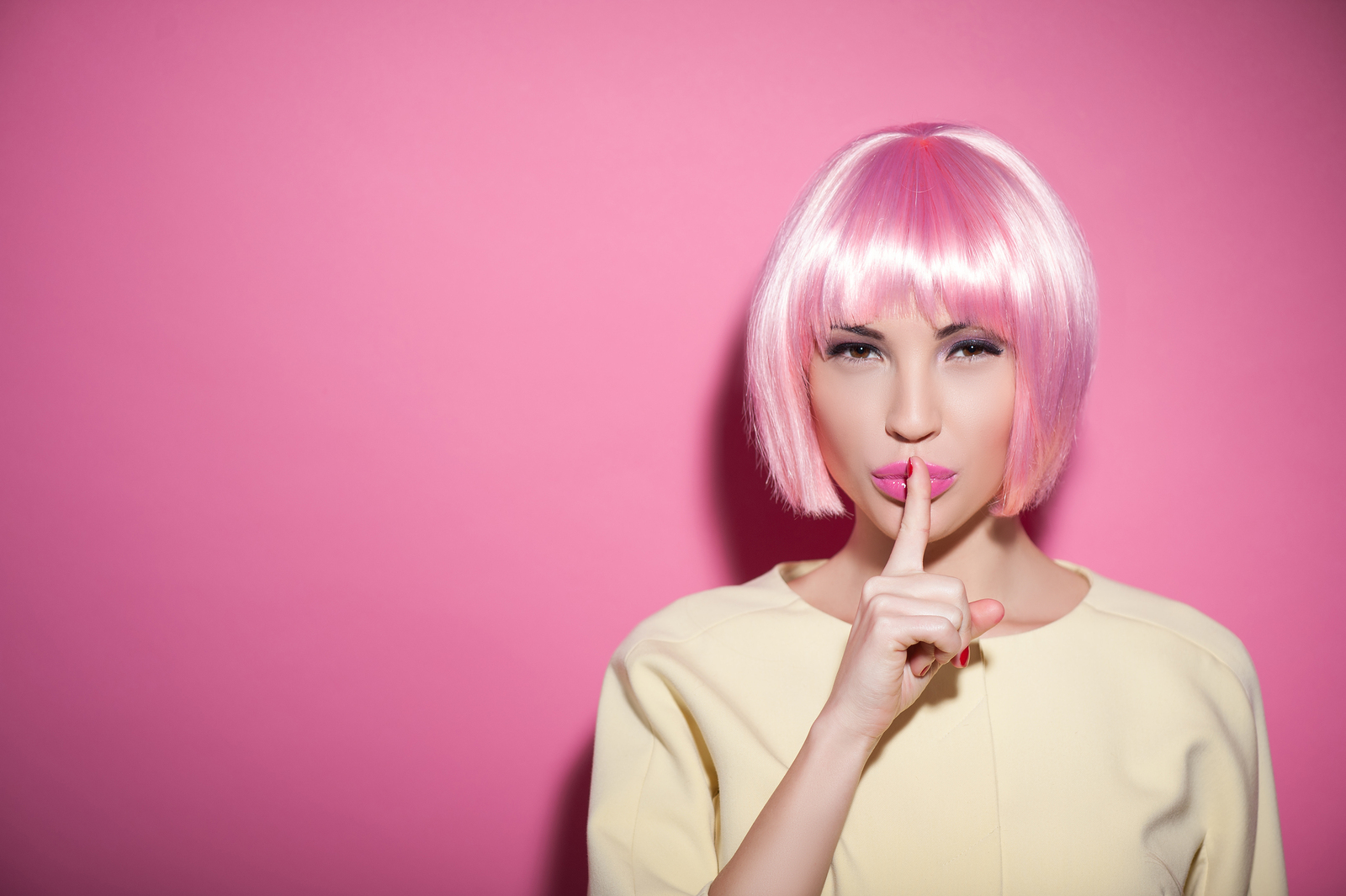 Save my secret. Portrait of attractive woman in a wig raising finger to her pink lips. She is standing and looking at camera mysteriously. Isolated and copy space in left side